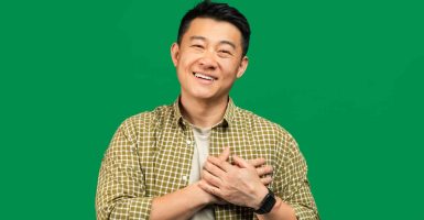Positive middle aged asian man praying and smiling at camera over green studio background, holding hands on his chest over heart, free space