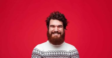 Handsome bearded guy in stylish warm sweater cheerfully smiling while standing on bright red background