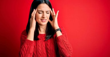 Young brunette woman with blue eyes wearing casual sweater over isolated red background with hand on headache because stress. Suffering migraine.