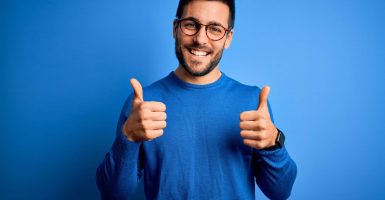 Young handsome man with beard wearing casual sweater and glasses over blue background success sign doing positive gesture with hand, thumbs up smiling and happy. Cheerful expression and winner gesture.